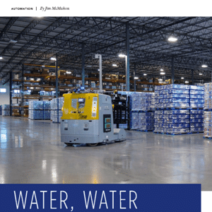 Automated forklift moves through a large warehouse with high shelves and stacked pallets of goods under bright overhead lights.