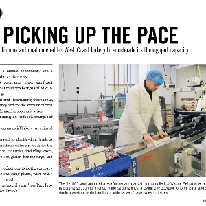 A bakery worker in a white coat and blue hairnet operates a packaging machine in a commercial bakery facility.